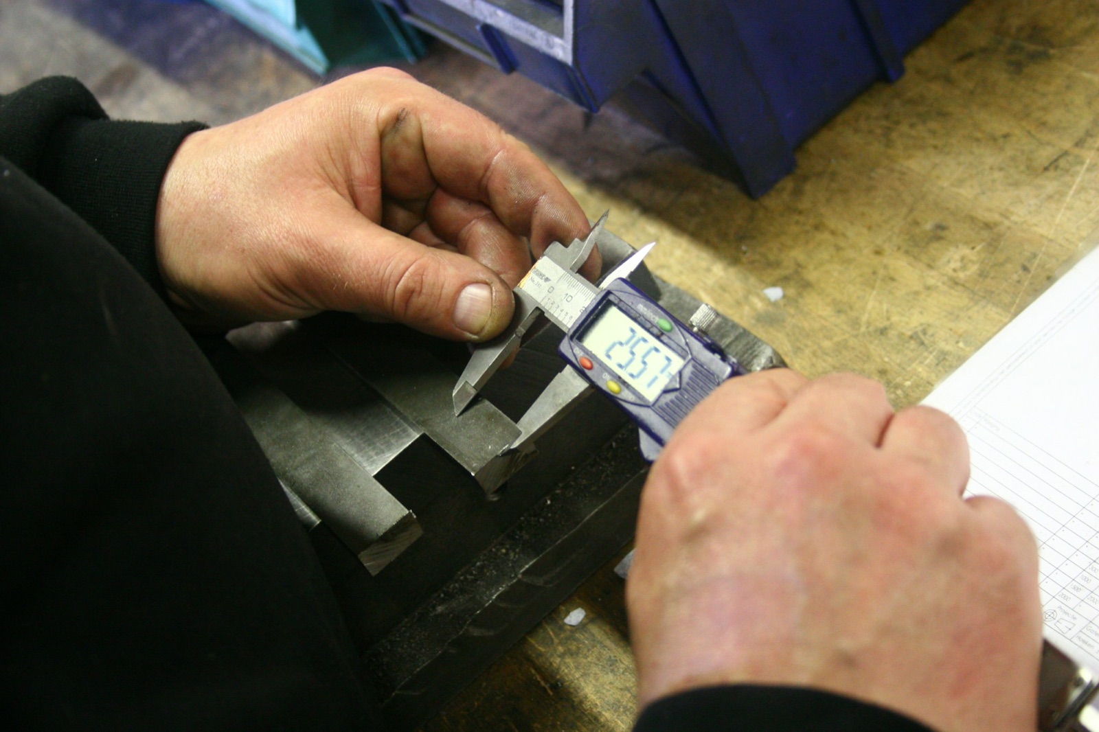 Inspector's hands measuring a metal part with a digital caliper on a workbench